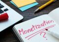An open notebook with "Monetization ?" written in red marker, placed next to a laptop, colorful sticky notes, and a yellow pen on a wooden desk.