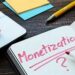 An open notebook with "Monetization ?" written in red marker, placed next to a laptop, colorful sticky notes, and a yellow pen on a wooden desk.