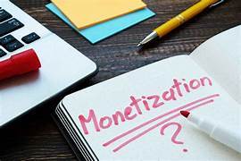 An open notebook with "Monetization ?" written in red marker, placed next to a laptop, colorful sticky notes, and a yellow pen on a wooden desk.