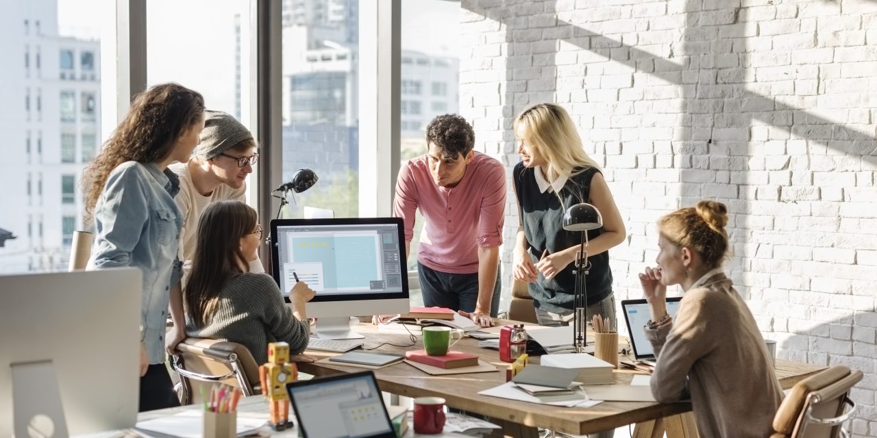 A high-angle shot of a dynamic team meeting in an open-plan office, focusing on a central computer screen, with natural light streaming in.